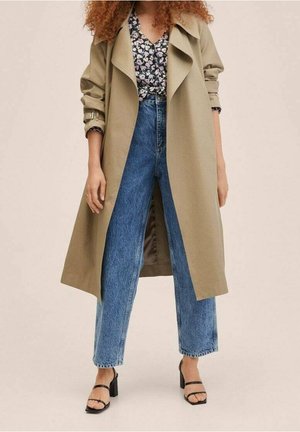 Beige Trenchcoat über einer schwarzen Bluse mit Blumenmuster, kombiniert mit hochtaillierten blauen Jeans und schwarzen Absatzsandalen.
