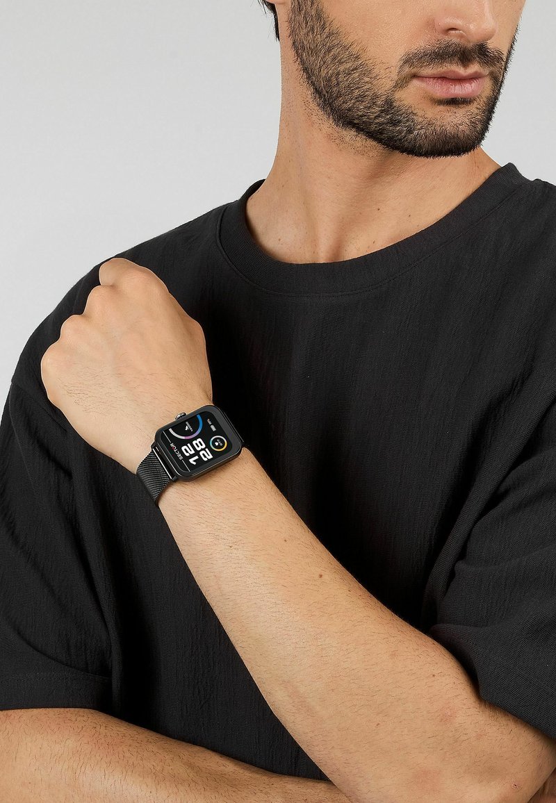 Orologio smartwatch nero con quadrante rettangolare, display digitale e cinturino in maglia, indossato su un polso. La maglietta è nera, a maniche corte e semplice.