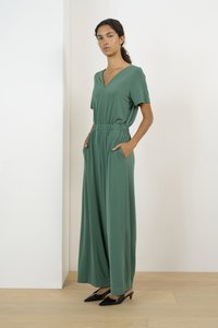 Mono verde de manga corta y con escote en V, con cintura elástica, bolsillos laterales y piernas anchas. Llevado con mule de tacón negro sobre un suelo de madera clara.