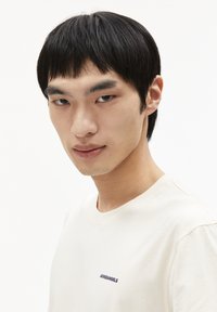 Beige T-shirt van zacht katoen, met een klein blauw logo op de borst, ronde halslijn en korte mouwen. Eenvoudig, minimalistisch ontwerp.