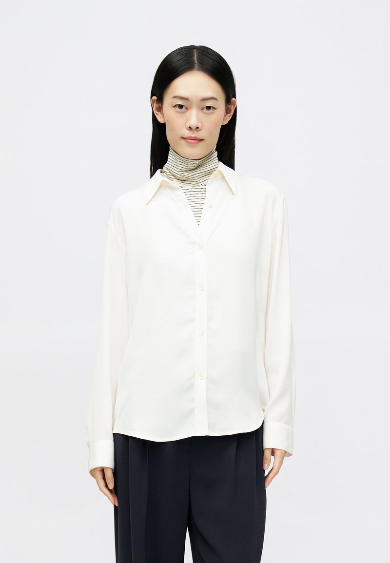 Mujer con cabello lacio y negro lleva una camisa blanca abotonada sobre un suéter de cuello alto a rayas blanco y negro y pantalones negros, de pie frente a un fondo blanco.