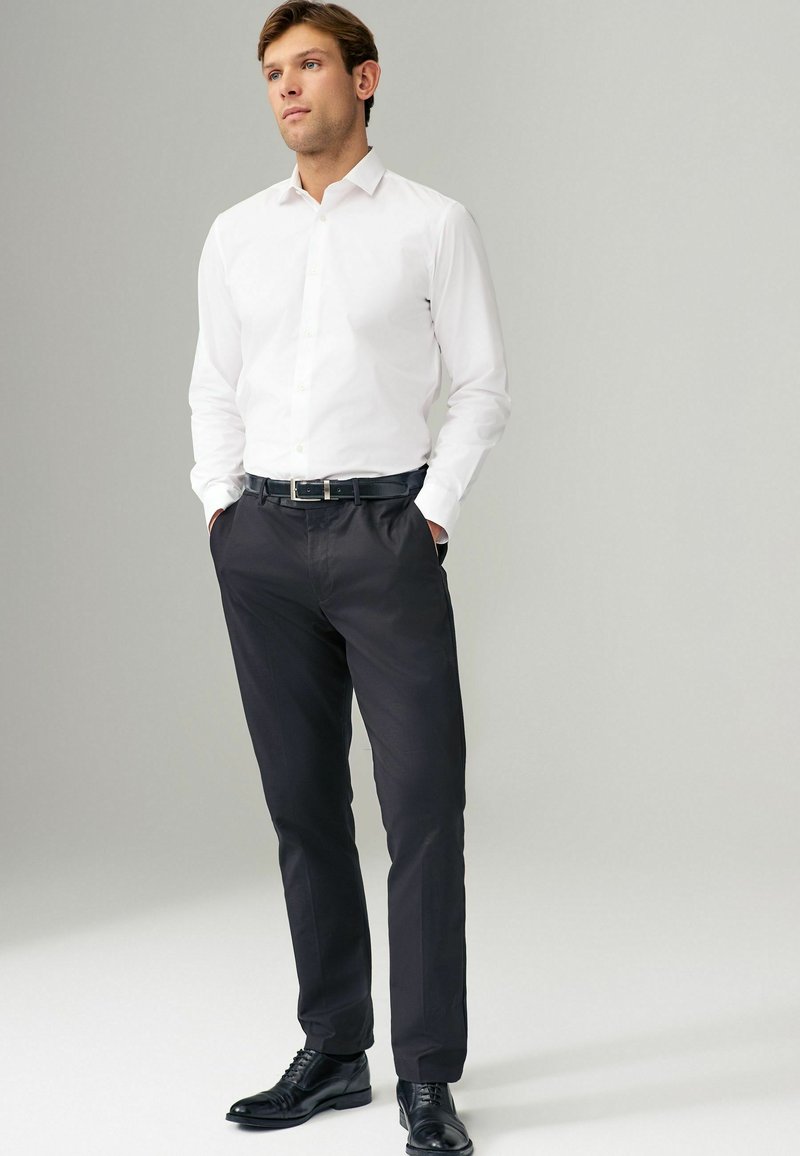 Chemise blanche à manches longues, rentrée dans un pantalon noir lisse, portée avec une ceinture noire et des chaussures noires cirées. Fond neutre.