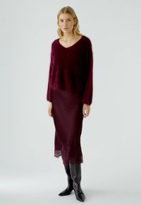 Bordeaux mohair blend trui met een V-hals en lange mouwen, gecombineerd met een bordeaux satijnen rok met kant aan de zoom. Zwarte kniehoge laarzen.