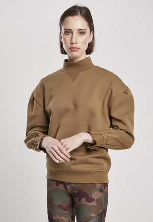 Brun sweatshirt med høj krave, rummelige ærmer og struktureret finish. Modellen parrer den med leggings i camouflage-mønster.
