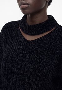 Gros plan sur une personne portant un pull en maille côtelée noire avec un col roulé épais roulé et une petite découpe en V au niveau du col.