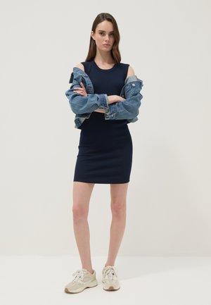 Jonge vrouw met lang bruin haar draagt een navy mouwloze jurk en een spijkerjasje dat van de schouders is afgeschoven, staat met gekruiste armen en witte sneakers.