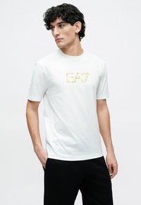 Λευκό βαμβακερό μπλουζάκι με κοντά μανίκια, που διαθέτει χρυσό λογότυπο "EA7" στο μπροστινό μέρος. Συνδυάζεται με μαύρο παντελόνι σε άνετη εφαρμογή.