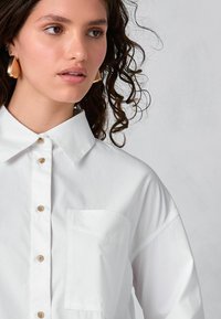 White collared shirt made of lightweight fabric, featuring a button front and a chest pocket. Notable details include tan buttons and rolled sleeves.