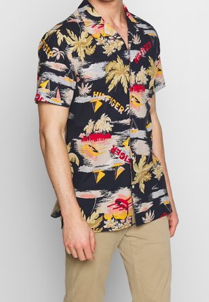 Man wearing short-sleeve black Hawaiian shirt with yellow palm trees, sailboats, red and yellow text, paired with beige pants.