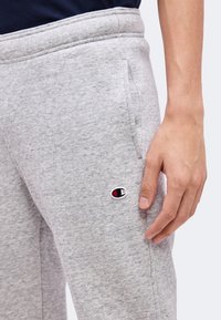 Zbliżenie na rękę osoby spoczywającą obok jasnoszarych dresów z małym logo Champion w pobliżu kieszeni.