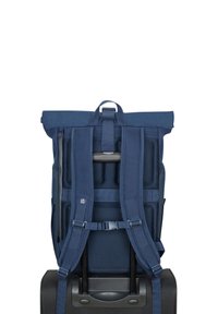 Sac à dos bleu marine avec un rouleau en haut, doté de bretelles rembourrées en mesh, de poches zippées et d'une poignée solide, reposant sur une valise à roulettes noire.