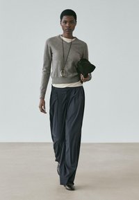 Grauer Pullover, weißes Unterhemd, marineblaue Faltenhose, schwarze Schuhe, hält eine kleine schwarze Tasche, mit einer langen Anhängerkette.