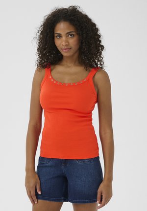 Femme aux cheveux bouclés portant un haut sans manches orange avec des œillets métalliques et un short en denim bleu foncé, debout devant un fond uni.