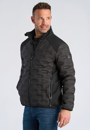 Mann trägt eine schwarze gesteppte Jacke mit Reißverschlusstaschen und blaue Jeans, schaut nach links vor einem schlichten Hintergrund.
