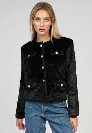 Femme portant une veste noire en fausse fourrure bouclée avec quatre poches et un jean bleu, debout devant un fond gris clair uni.