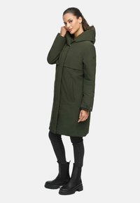 Manteau vert, longueur genou, avec une capuche, un rabat avant oversize et des poches latérales. Associé à un jean noir et des bottes noires imposantes.