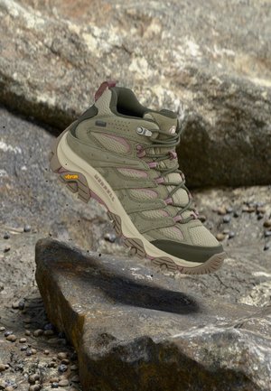 Botte de randonnée avec semelle épaisse et lacets suspendue au-dessus d'un terrain rocheux, mettant en valeur l'adhérence et l'utilisation en extérieur.