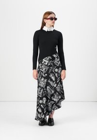 Un model stă în fața unui fundal alb, purtând un pulover negru ajustat, o fustă cu model alb-negru și ochelari de soare, evidențiind un look elegant.