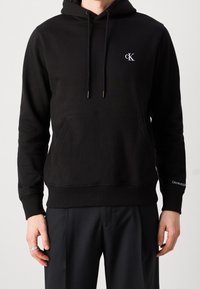 Sudadera negra con capucha que incluye un bolsillo frontal, capucha ajustable con cordón y un pequeño logo blanco "CK" en el pecho. Confeccionada en tejido suave y con textura.