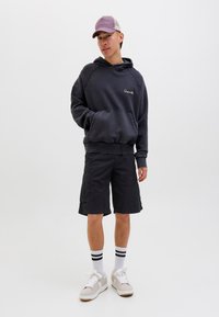 Šedá oversized mikina s kapucí a kapsou, černé cargo šortky, bílé tenisky s hnědými akcenty a proužkované bílé ponožky. Obsahuje čepici s fialovým kšiltem.