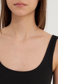 Haut noir côtelé avec un large décolleté en scoop. Texture lisse, fines bretelles et coupe ajustée. Teint de peau visible en arrière-plan.