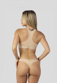 Ensemble brassière de sport et tanga beige en tissu doux. Présente un design dos nageur et une bande logo à la taille. Texture lisse, détails minimaux.