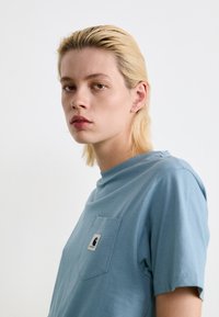 Jeune personne aux cheveux blonds courts portant un t-shirt Carhartt bleu clair, faisant face à l'avant avec une expression neutre sur un fond blanc.