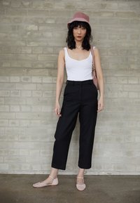 Femme debout contre un mur en briques portant un chapeau rose, un haut blanc sans manches, un pantalon noir coupé, des chaussures plates roses, et portant un sac à main rose.