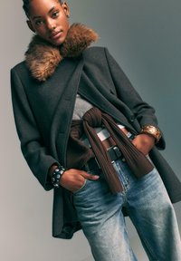 Donna che indossa un cappotto grigio scuro con colletto di pelliccia, top grigio chiaro, maglione marrone legato in vita, jeans azzurri e due braccialetti con borchie in metallo, mani in tasca.