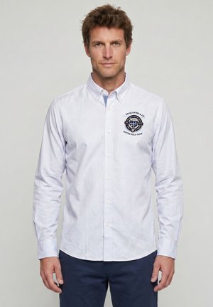 Hombre vestido con una camisa blanca de botones con el logo "Valecuatro & Co United Polo Team" en el pecho, combinado con pantalones oscuros.