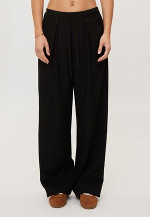 Personne portant un pantalon noir ample avec taille à cordon et des chaussures marron style mocassin, debout devant un fond clair uni.