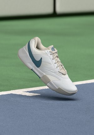 Weißer und beiger Nike-Sneaker mit blauem Swoosh, der über einer blau-grünen Sportplatzoberfläche schwebt.