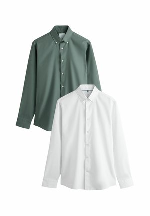 Jeune homme debout, les mains dans les poches, portant une chemise turquoise à boutons, un pantalon beige, une ceinture marron et des chaussures noires sur fond uni.