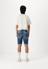Pantalones cortos de mezclilla azul con dobladillos enrollados, usados con una camiseta blanca y calcetines hasta el tobillo. Se observan los bolsillos y una etiqueta negra en la cinturilla.