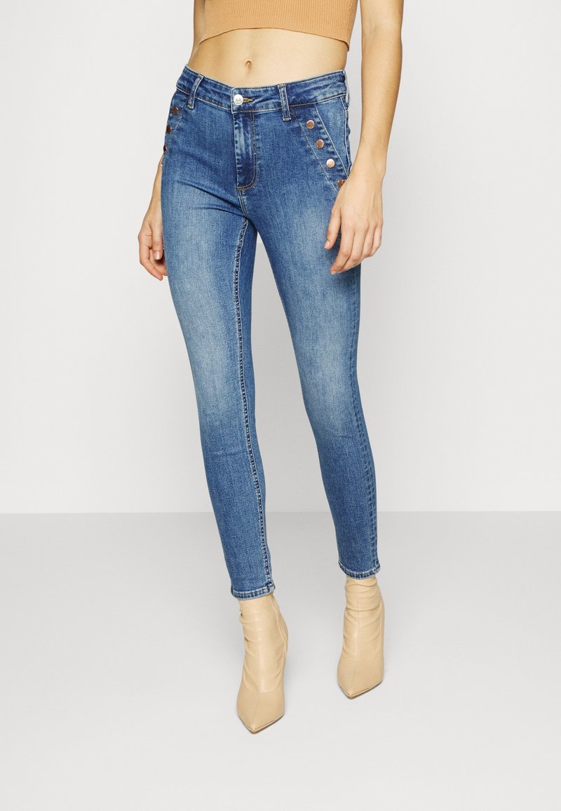 Femme portant un jean skinny bleu délavé taille haute avec des détails de boutons métalliques et des bottines beiges à bout pointu, debout sur un sol blanc.