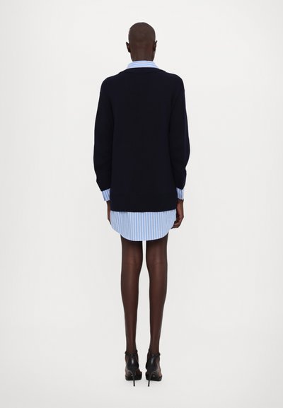 Marineblaues Strickpullover, der über einem hellblauen gestreiften Hemdkleid getragen wird. Der Pullover hat gerippte Bündchen und Saum, eine lässige Passform und lange Ärmel.