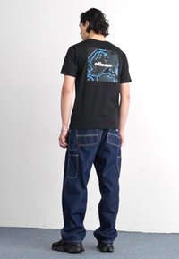 Juodas medvilnės marškinėlis su mėlynu abstrakčiu piešiniu ir "ellesse" logotipu ant nugaros, derinamas su laisvo kirpimo mėlynomis džinsinėmis kargo kelnėmis.