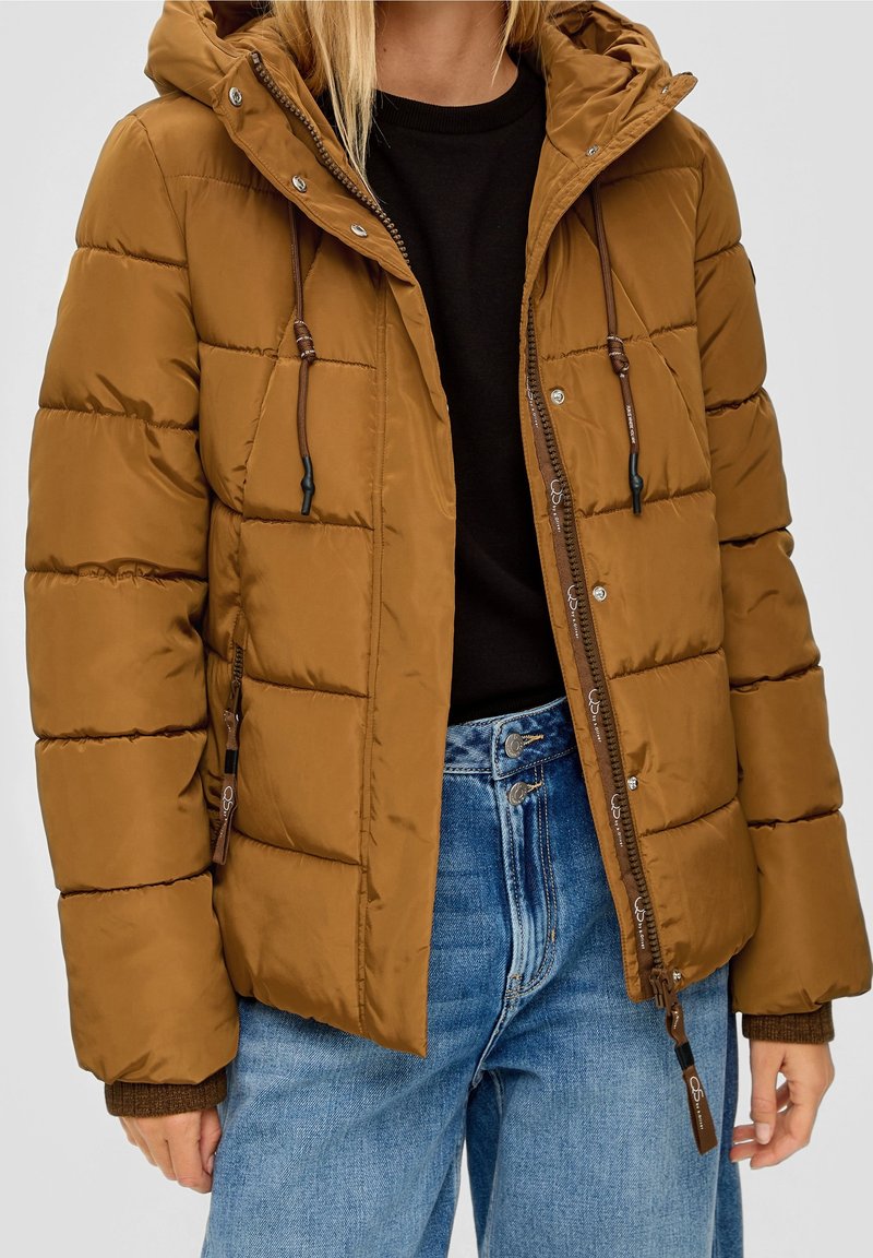 Femme portant une veste matelassée marron avec des poches zippées par-dessus une chemise noire et un jean bleu taille haute.