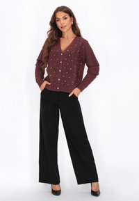 Femeie îmbrăcată într-o cardigan tricotat de culoare vișinie, cu detalii din perle, și pantaloni negri cu croială largă, pozând cu mâinile în buzunare și pantofi cu toc negri.