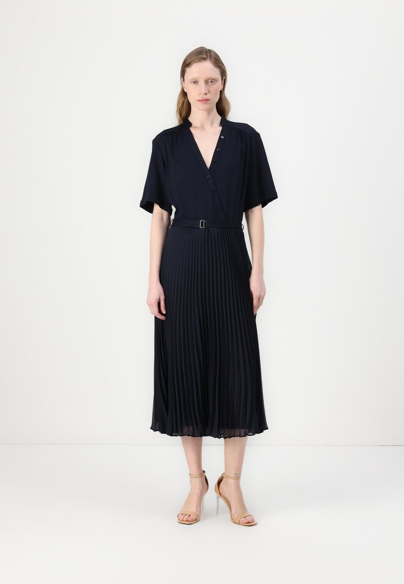 Navy pleated dress with a V-neck, short sleeves, and a belted waist. Smooth texture with a flowing silhouette extending to the mid-calf.
