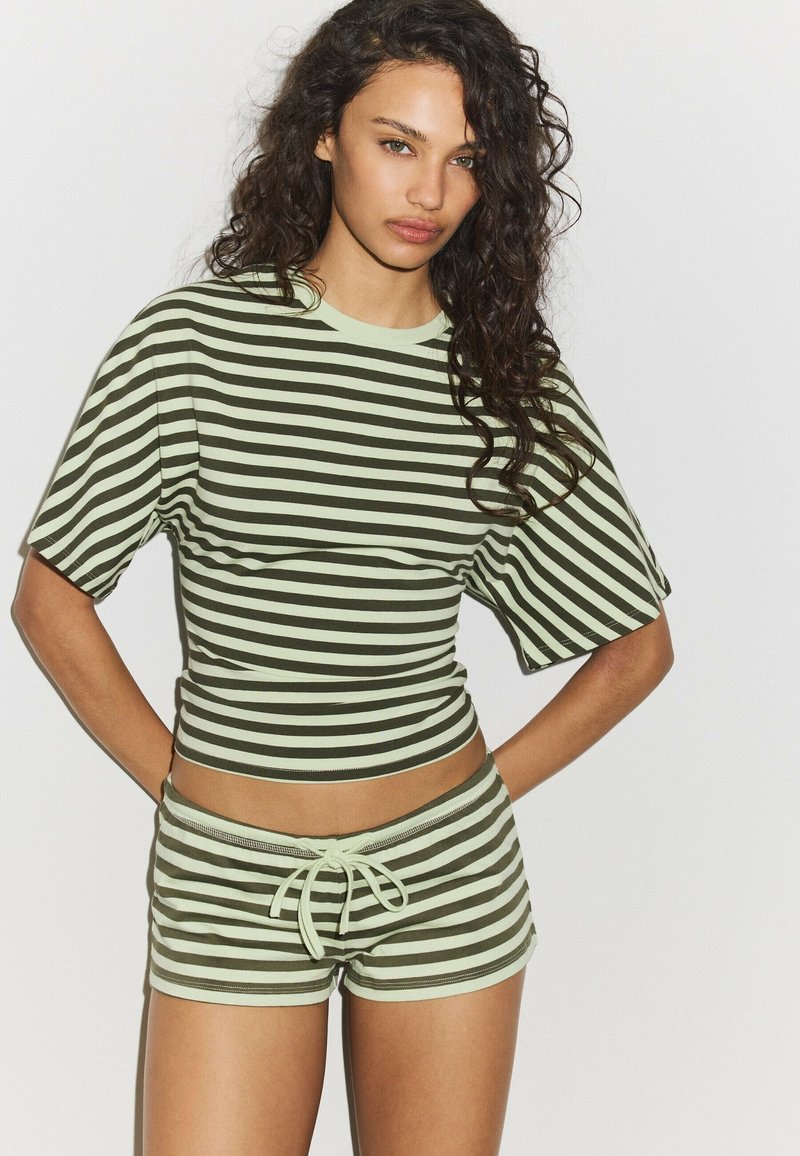 Jeune femme aux cheveux bouclés porte un crop top rayé horizontal vert et blanc assorti ainsi que des shorts à cordon, debout devant un fond uni.