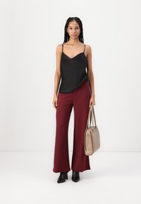 Zwarte satijnen camisole met dunne bandjes, gecombineerd met bordeauxrode wijde broeken. Model houdt een lichtbeige handtas vast en draagt zwarte hakboots.