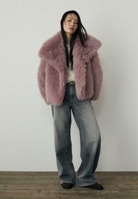 Puha roza umetna krznena jakna s širokimi reverji, v kombinaciji s svetlo sivo širokimi hlačami in temno rjavimi čevlji, postavljena pred nevtralno ozadje.