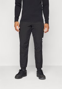 Juodos sportinės kelnės su siauru apipavidalinimu, pagamintos iš lengvo audinio, turinčios užtrauktuku užsegamas kišenes ir baltą logotipo akcentą kairėje šlaunies pusėje.