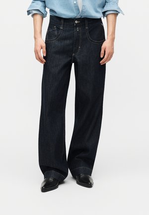 Άτομο που φοράει φαρδιά σκούρα τζιν παντελόνια, γαλάζιο πουκάμισο με ρεβέρ, και μαύρα μυτερά παπούτσια, στέκεται μπροστά σε απλό φόντο.