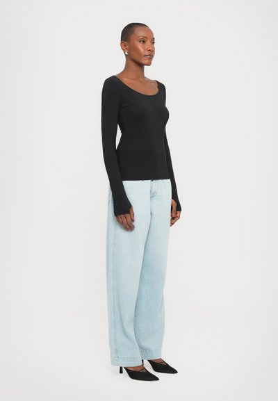Maglietta nera a maniche lunghe con scollatura a girocollo, abbinata a jeans in denim blu chiaro a gamba larga e tacchi a punta neri, su uno sfondo bianco.