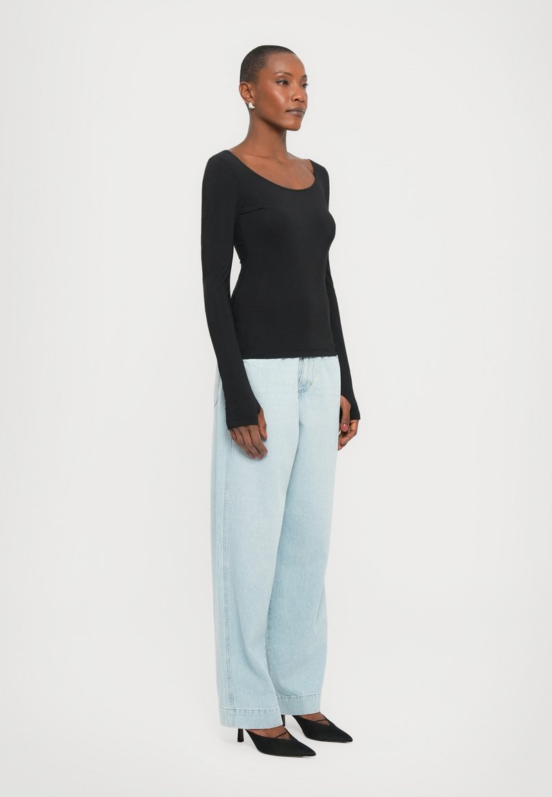 Un haut noir à manches longues avec un décolleté en ronde, associé à un jean en denim à jambes larges bleu clair et des escarpins noirs à bout pointu, sur un fond blanc.