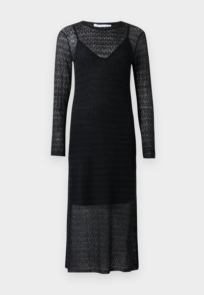 Robe noire en dentelle avec des manches longues, décolleté en V, motif texturé et coupe mi-longue. Comprend une partie inférieure fluide et légèrement transparente.