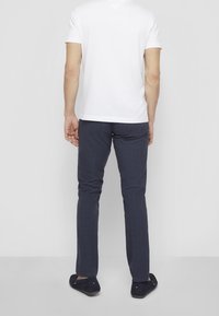 Muž v bílém krátkém rukávu, tmavě modrých kostkovaných kalhotách a tmavě modrých polobotkách stojící čelem ke bílému zdi.