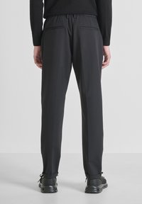 Persona che indossa pantaloni neri su misura e sneakers nere, in piedi con le mani rilassate lungo i fianchi, vista da dietro contro uno sfondo neutro.